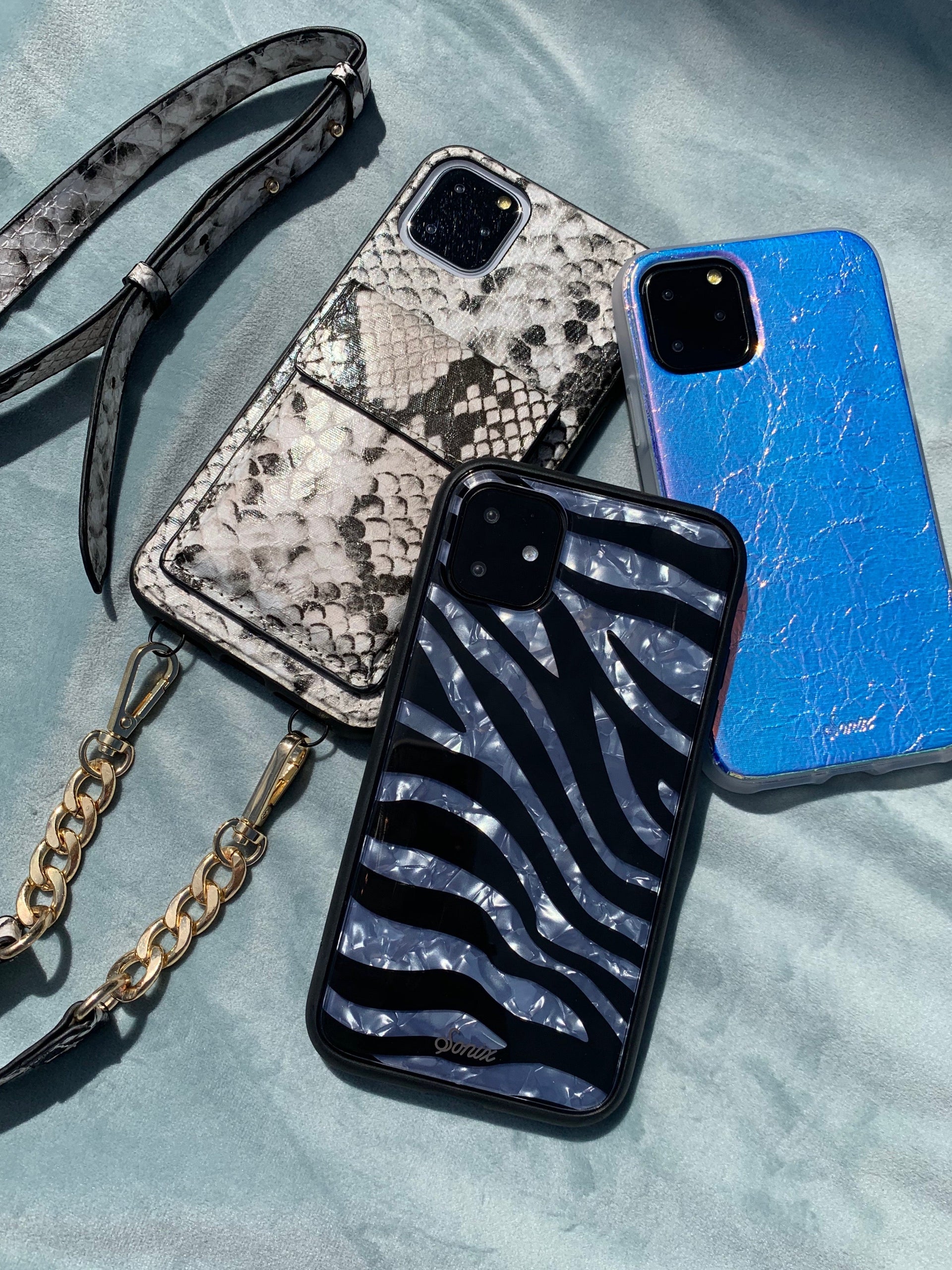 Flat lay of three Sonix iPhone cases showing zebra print, silver snakeskin wristlet, and iridescent blue finishes on pale blue fabric