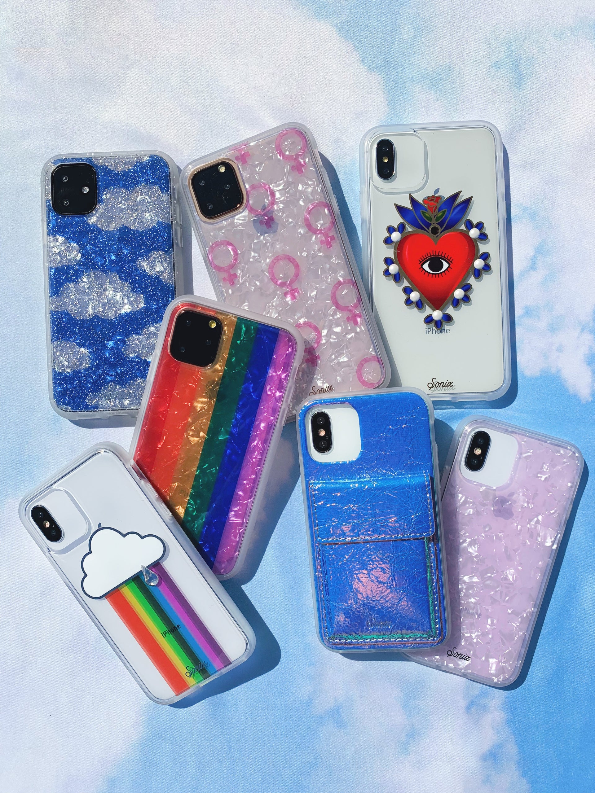 Overhead view of seven iPhone cases showing glitter, rainbow stripes, pearlescent shell, and a blue card pocket on cloud background