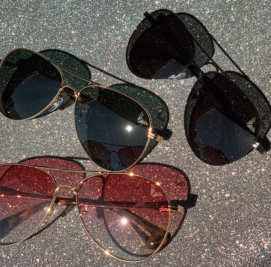 Flat lay of three Lodi aviator sunglasses showing gold frames and dark, black, and red lenses on a glittery silver background