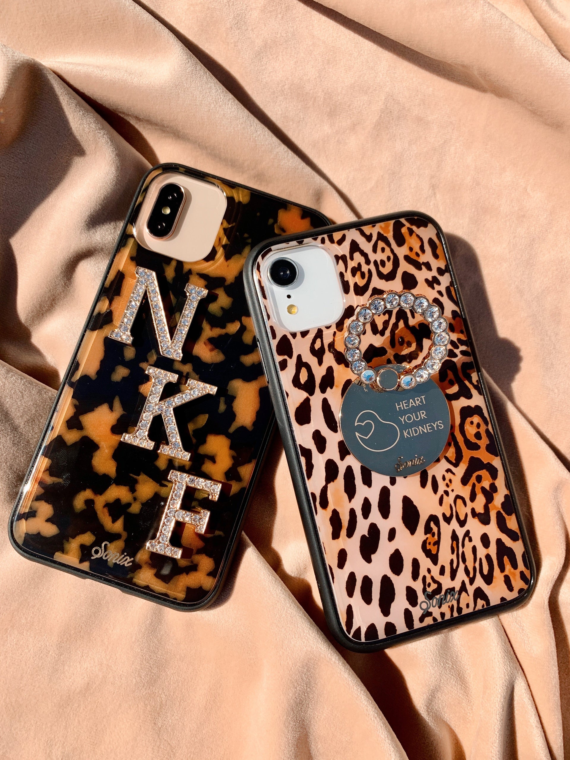 Angled view of two Sonix iPhone cases showing tortoise and leopard prints, rhinestone initials, and a crystal ring grip reading Heart Your Kidneys on pink satin