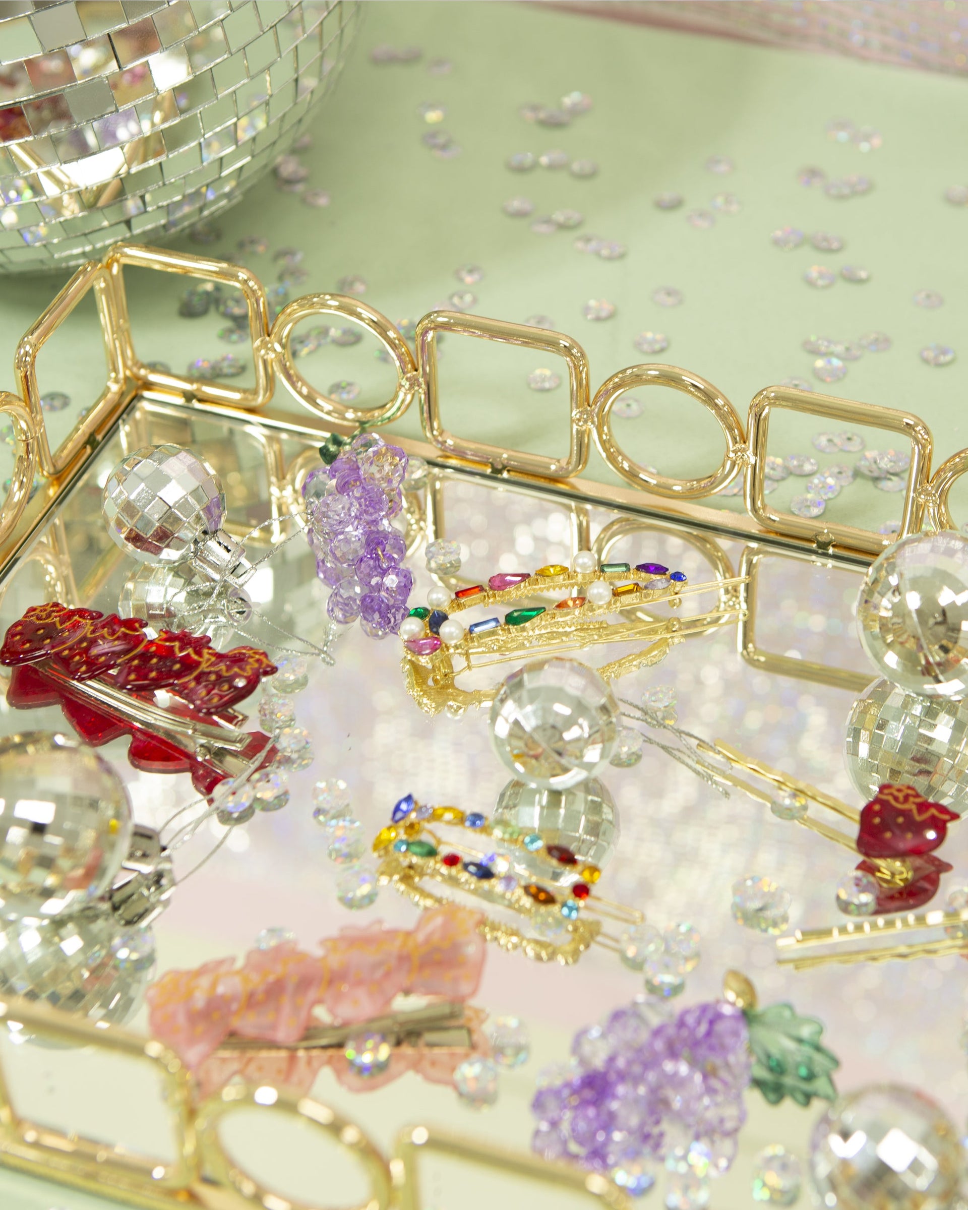 Decorative hair clips and mirror ornaments on a gold mirrored tray with scattered sequins on a pale green background