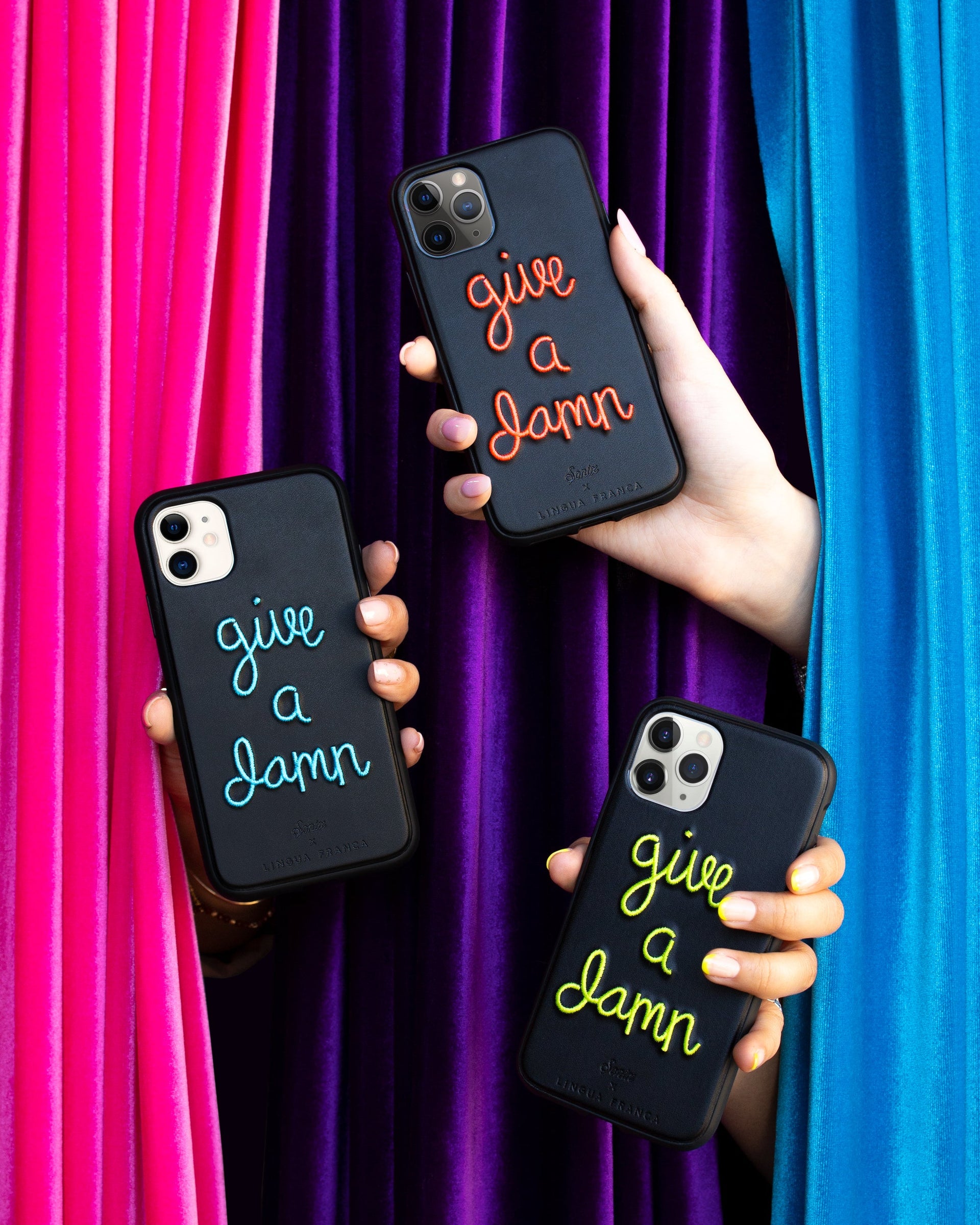 Angled view of iPhone case showcasing orange embroidered “give a damn” slogan with pink, purple and teal velvet curtain background