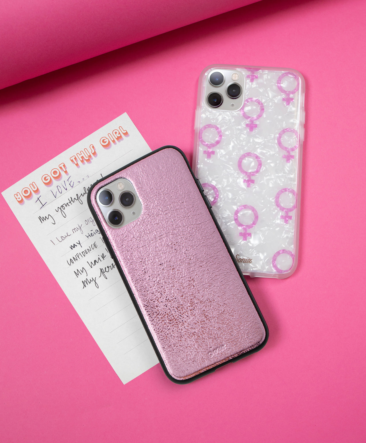 Top-down view of two iPhone cases: metallic pink textured case and clear pearlescent case printed with pink female symbols on a pink background