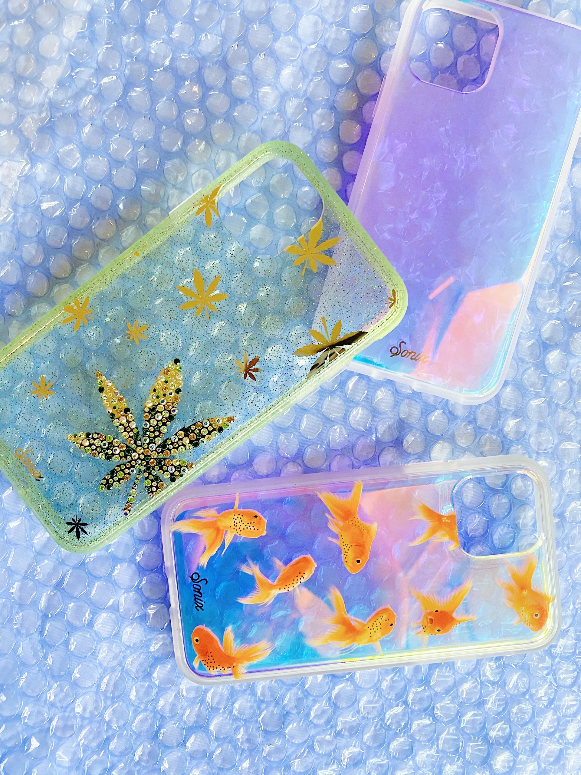 Angled view of three iPhone cases: jeweled green cannabis-leaf, iridescent purple, and goldfish-print on blue bubble wrap