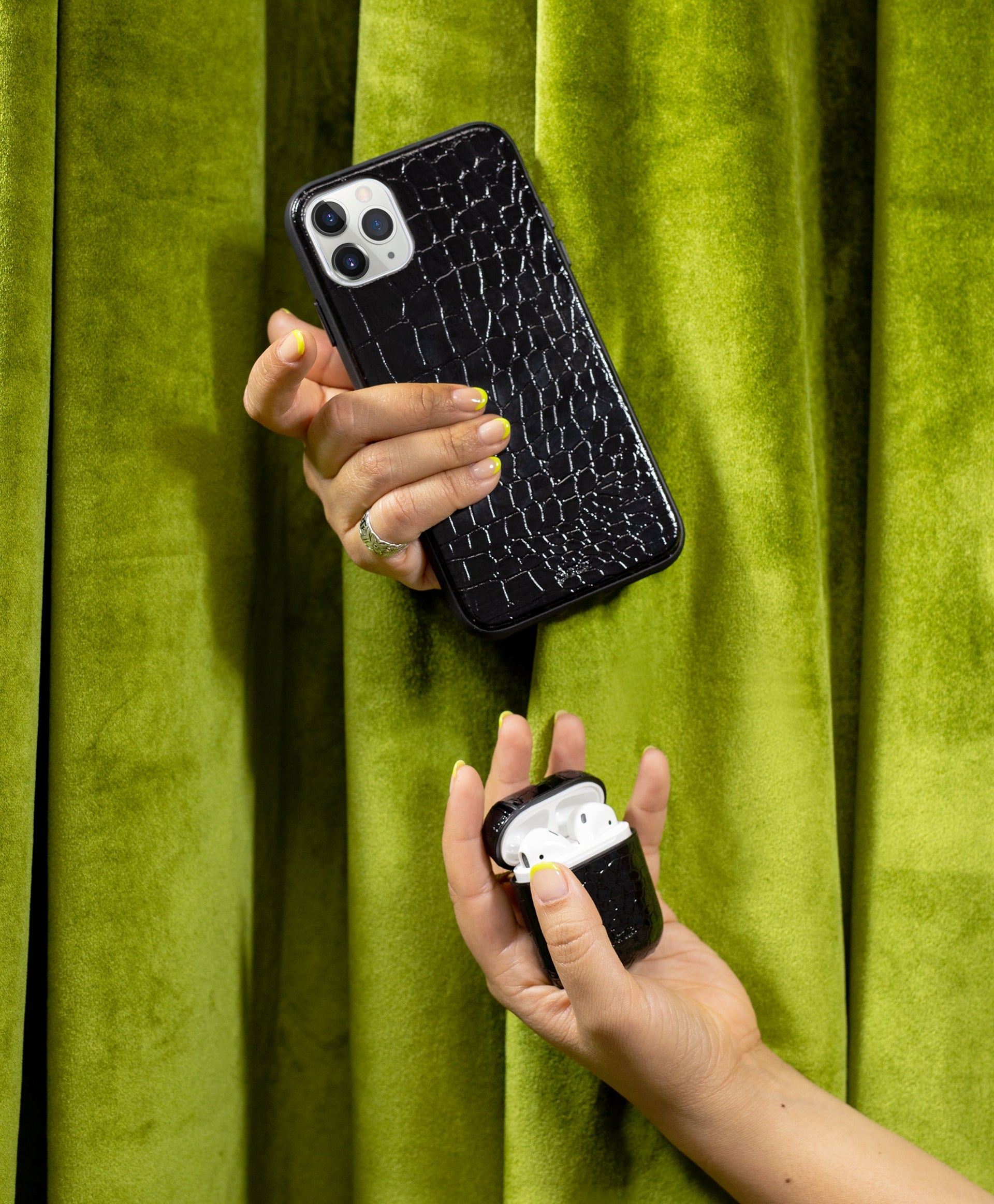 Angled back view of Onyx Croc iPhone case and matching AirPods case in hand showing shiny faux croc texture on green curtain