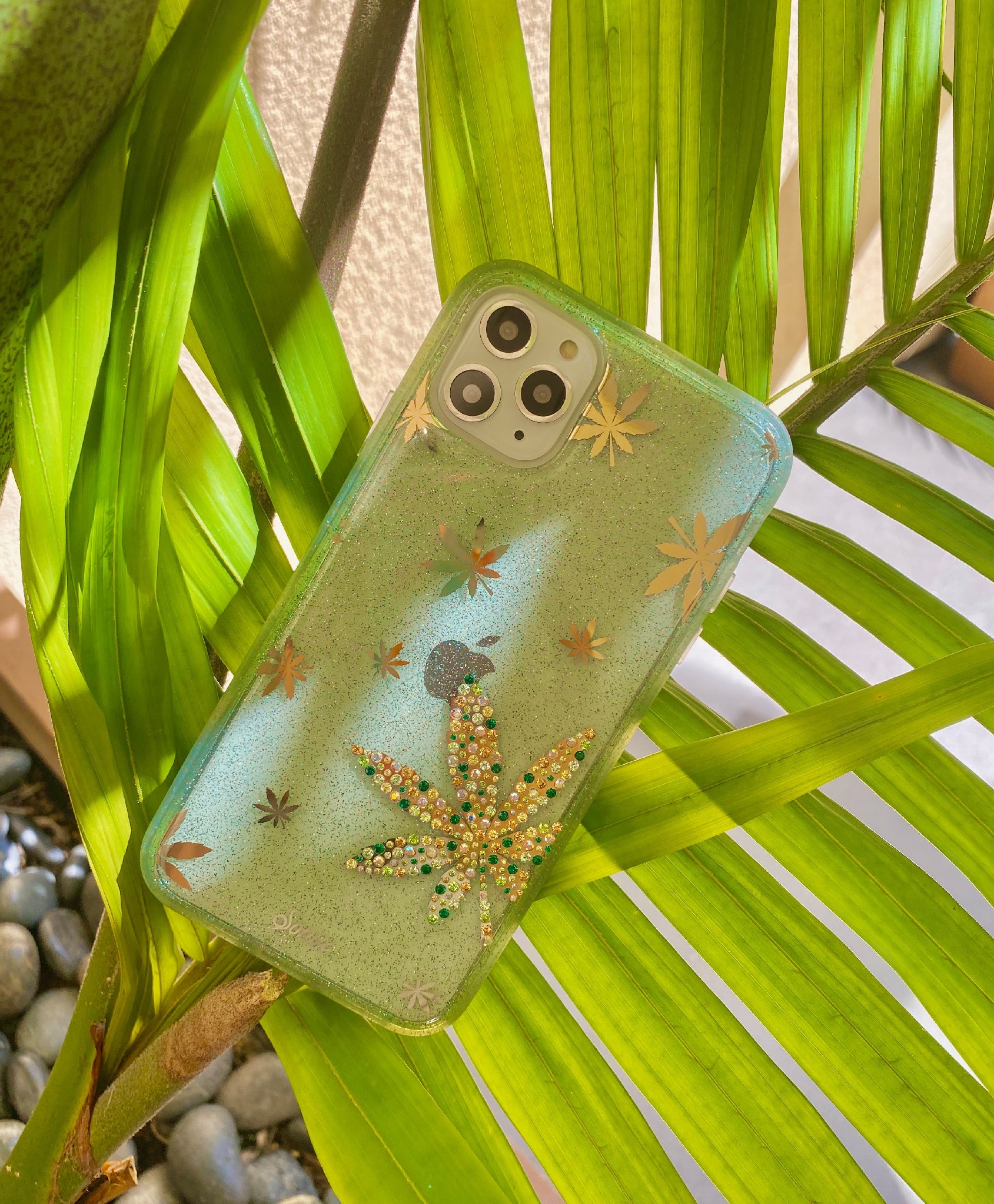 Angled close-up of Mary Jane glitter iPhone case with gold foil and green rhinestone leaf design and tinted green glitter bumper on palm leaves