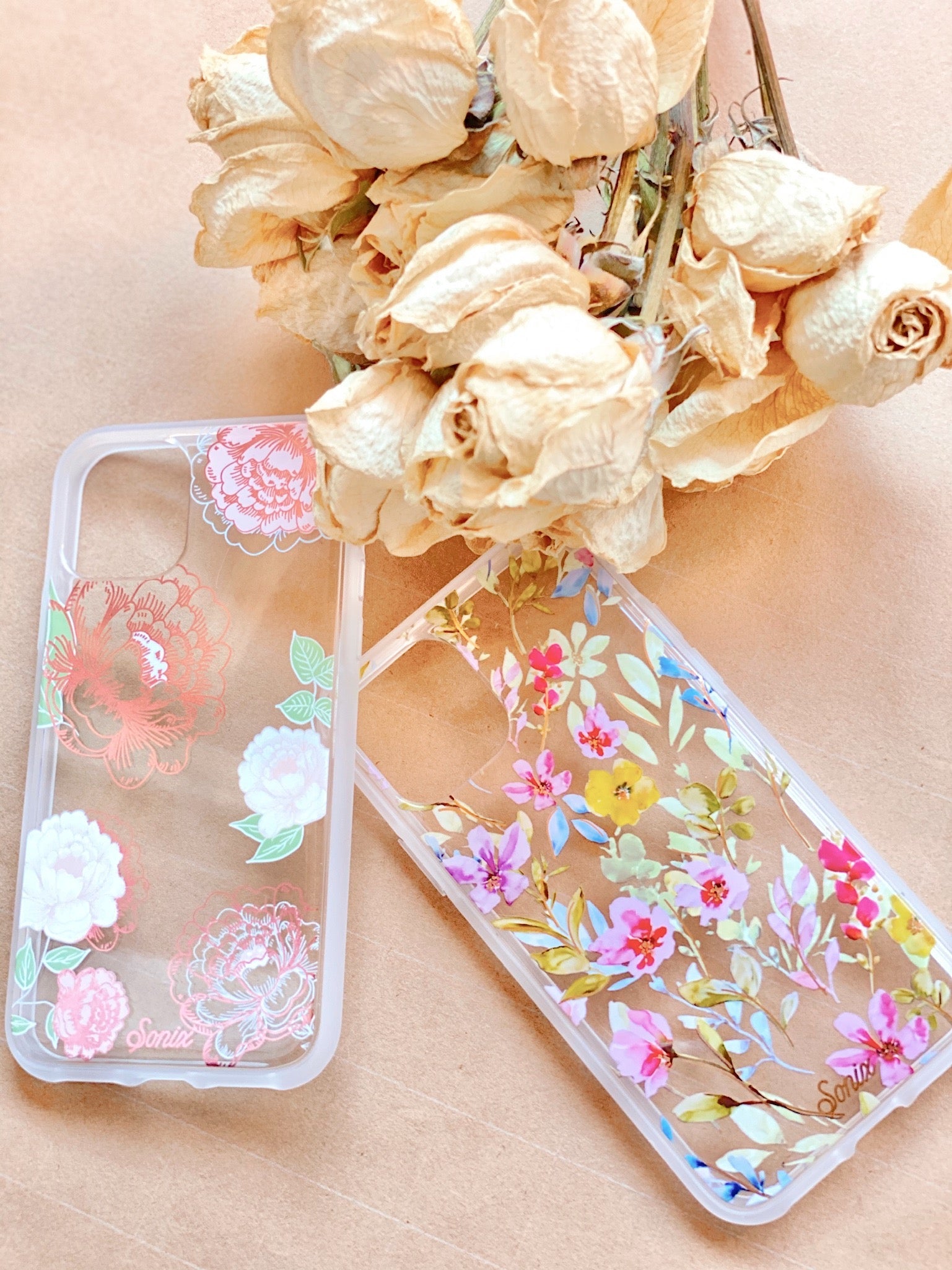 Flat lay of two clear iPhone cases with pink and multicolor floral prints and gold-foiled accents on a peach background with dried roses