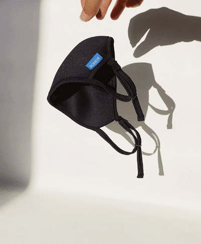 Angled view of a black contoured face mask showcasing adjustable ear loops, held above a white surface and casting a shadow