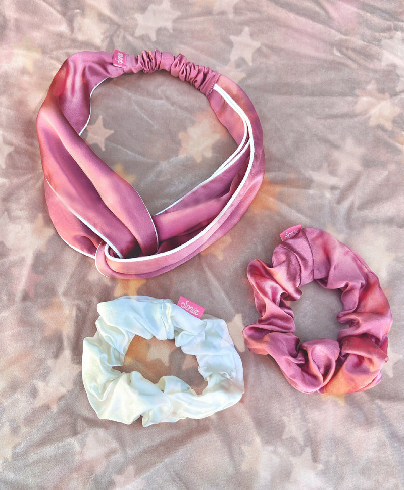Top-down view of a twist knot headband and two scrunchies in dusty mauve and cream 100% organic mulberry silk on a star-patterned pink cloth