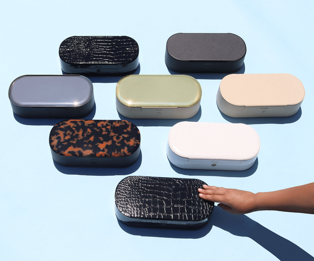 Overhead view of UV sanitizing boxes showcasing assorted finishes with a hand touching one on a light blue surface