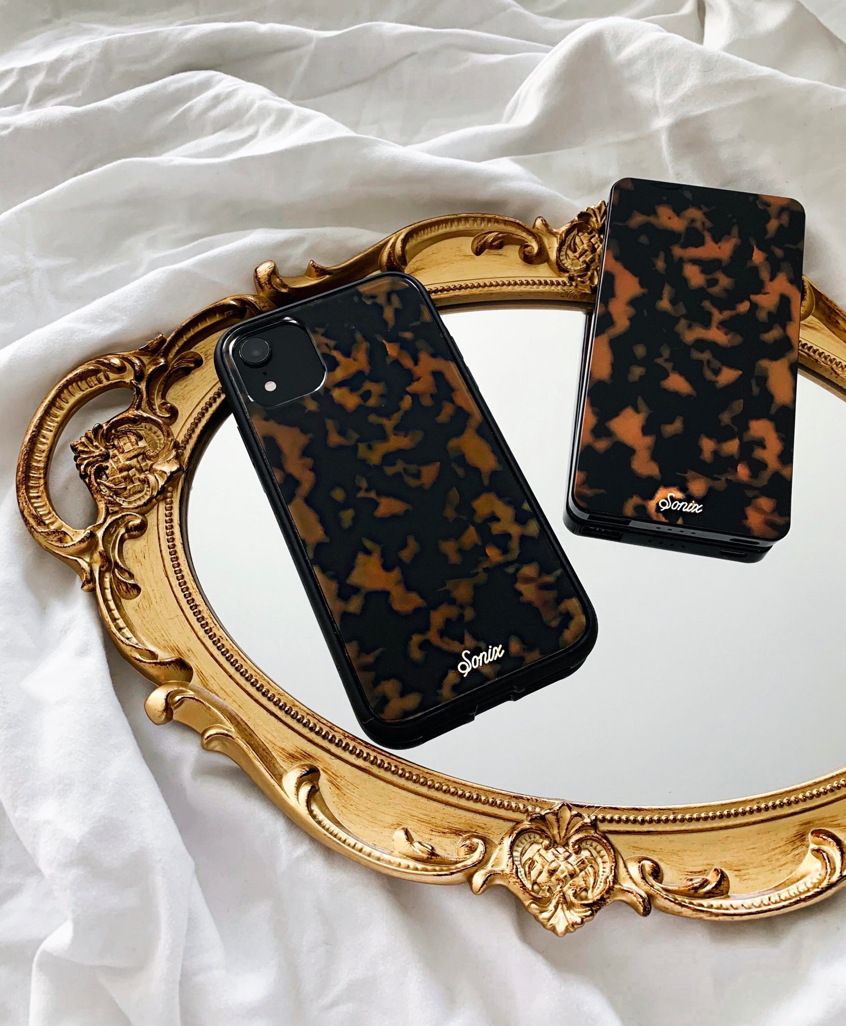 Angled view of Brown Tort iPhone cases with glossy tortoiseshell pattern and Sonix logo on a gold mirror over white sheet