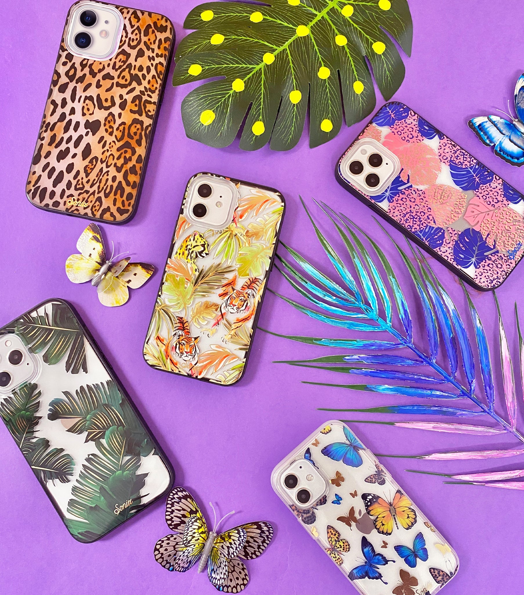 Several iPhone cases arranged on a purple background with tropical leaves, butterflies, and colorful patterns