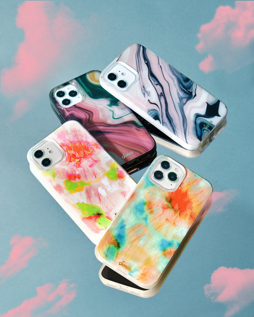 Angled flat-lay of four iPhone cases showcasing marbled and watercolor patterns against a blue background with pink cloud accents