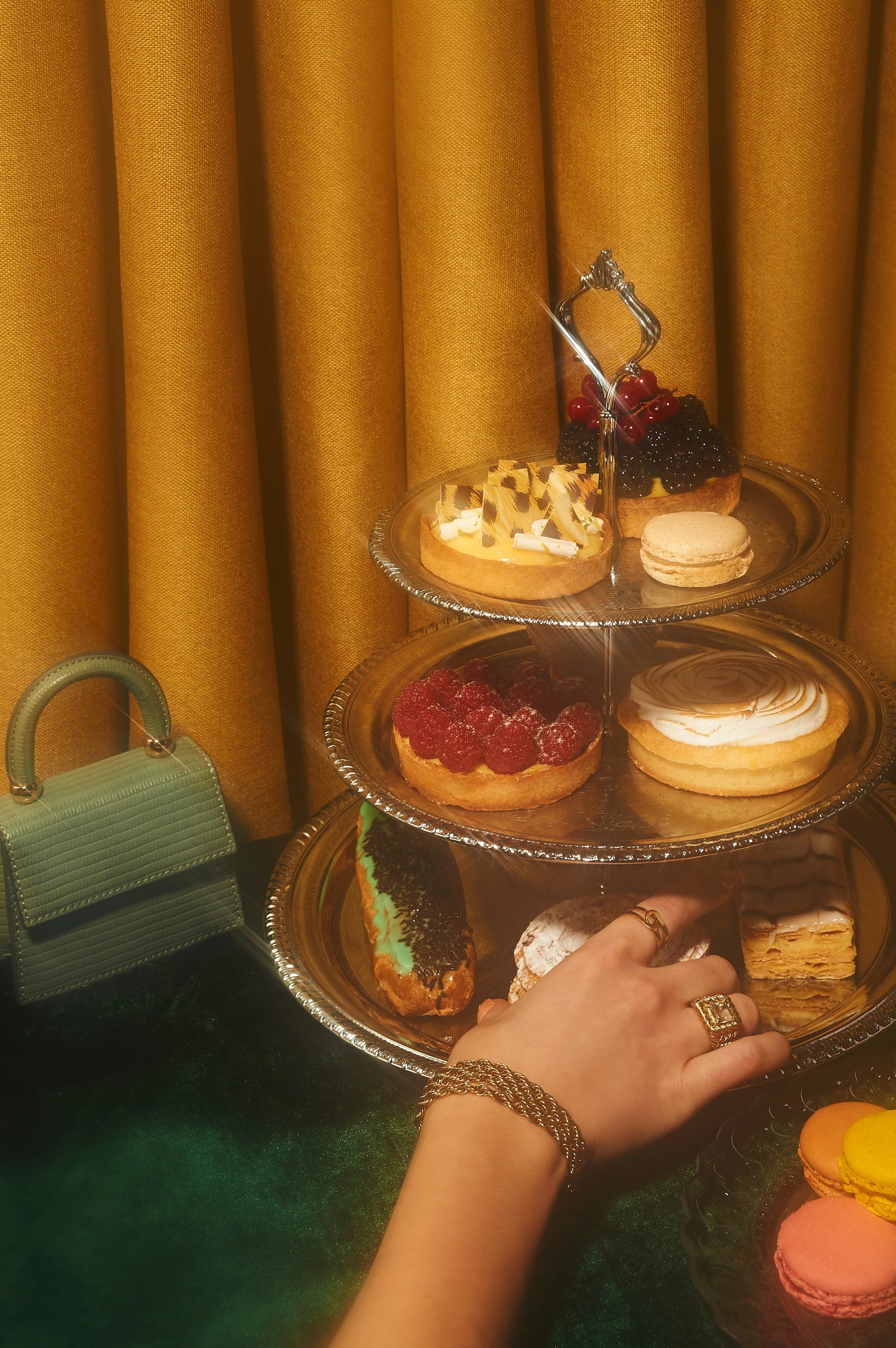 Hand reaching for pastries on a three-tiered silver tray beside macarons and a small green handbag against mustard-yellow curtains