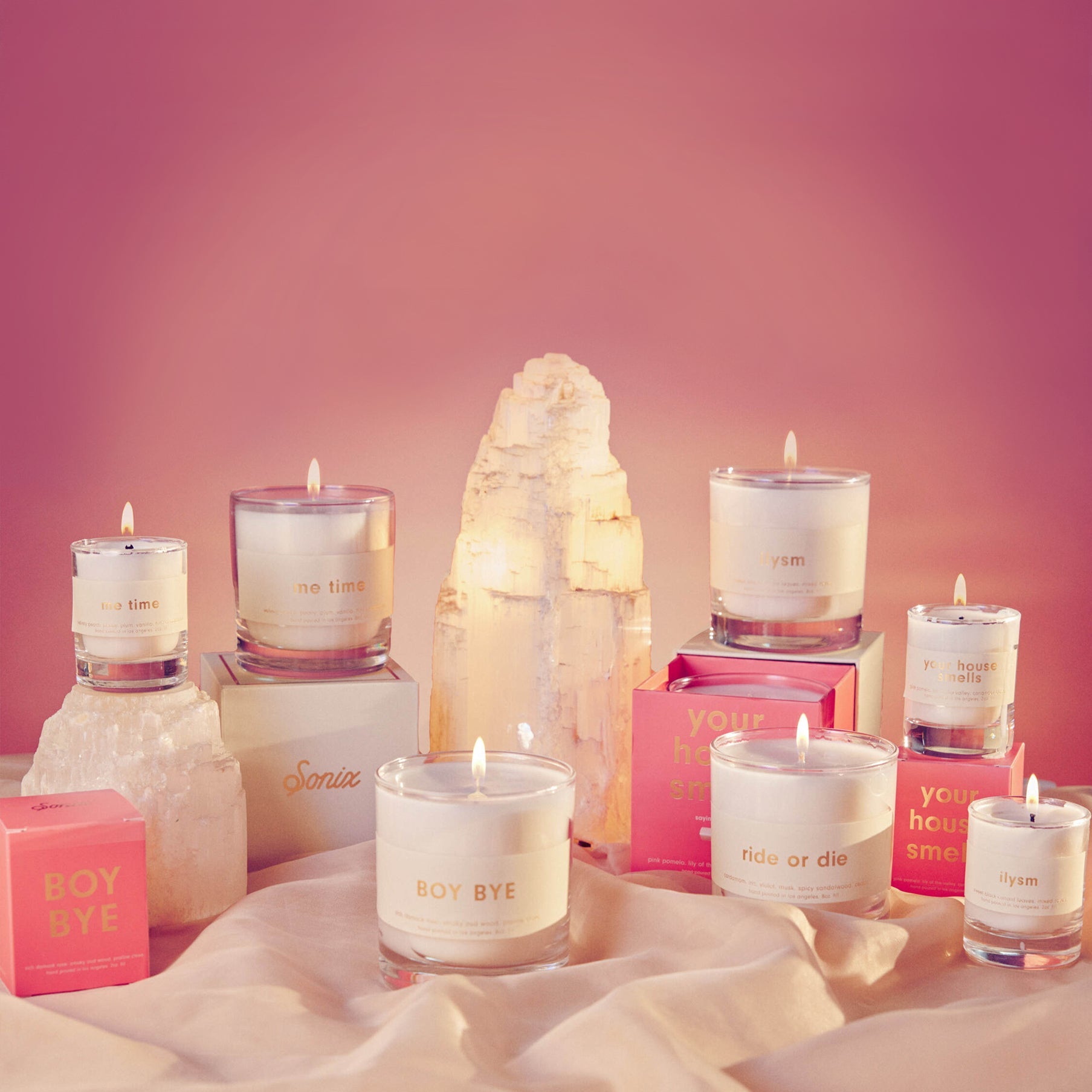 Front view of lit scented candles in clear glass jars labeled Boy Bye and ride or die, arranged with pink boxes and a selenite crystal on a pink gradient background