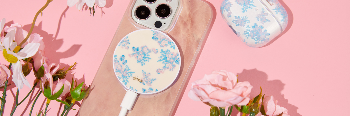 iPhone with a floral MagSafe charger attached, matching floral AirPods case, and pink roses on a pink background