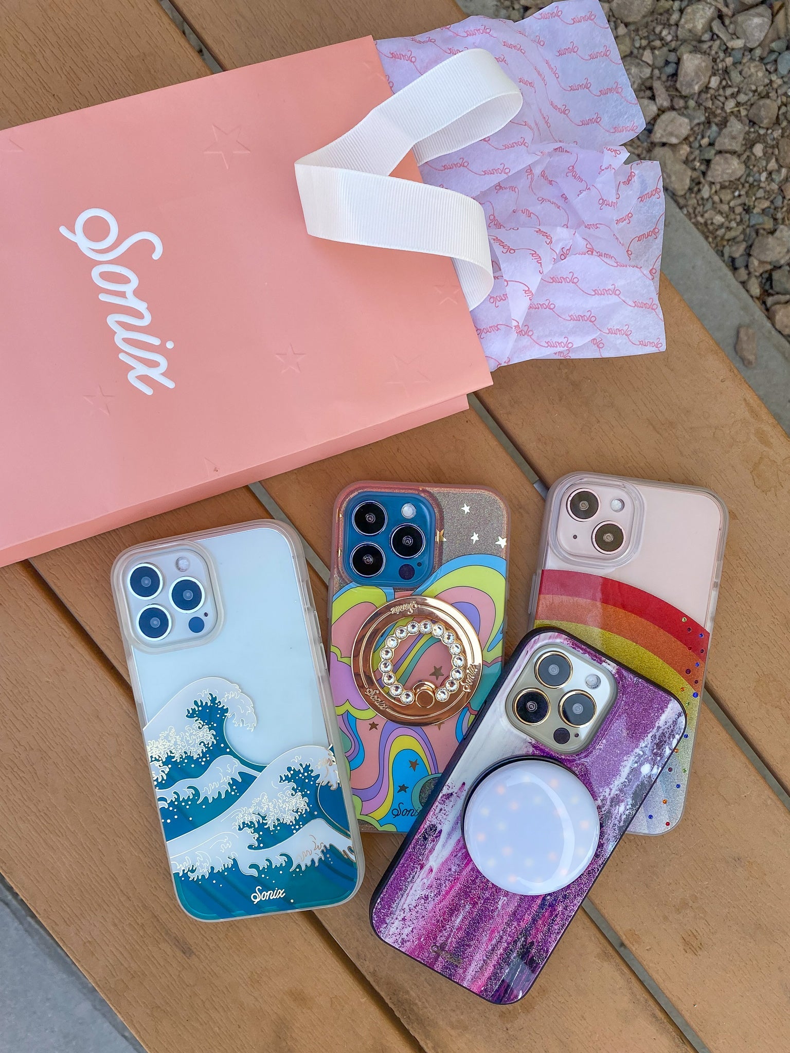 Flat lay of Sonix iPhone cases with colorful prints, rhinestone ring and pop out light on a wooden table with a pink Sonix bag