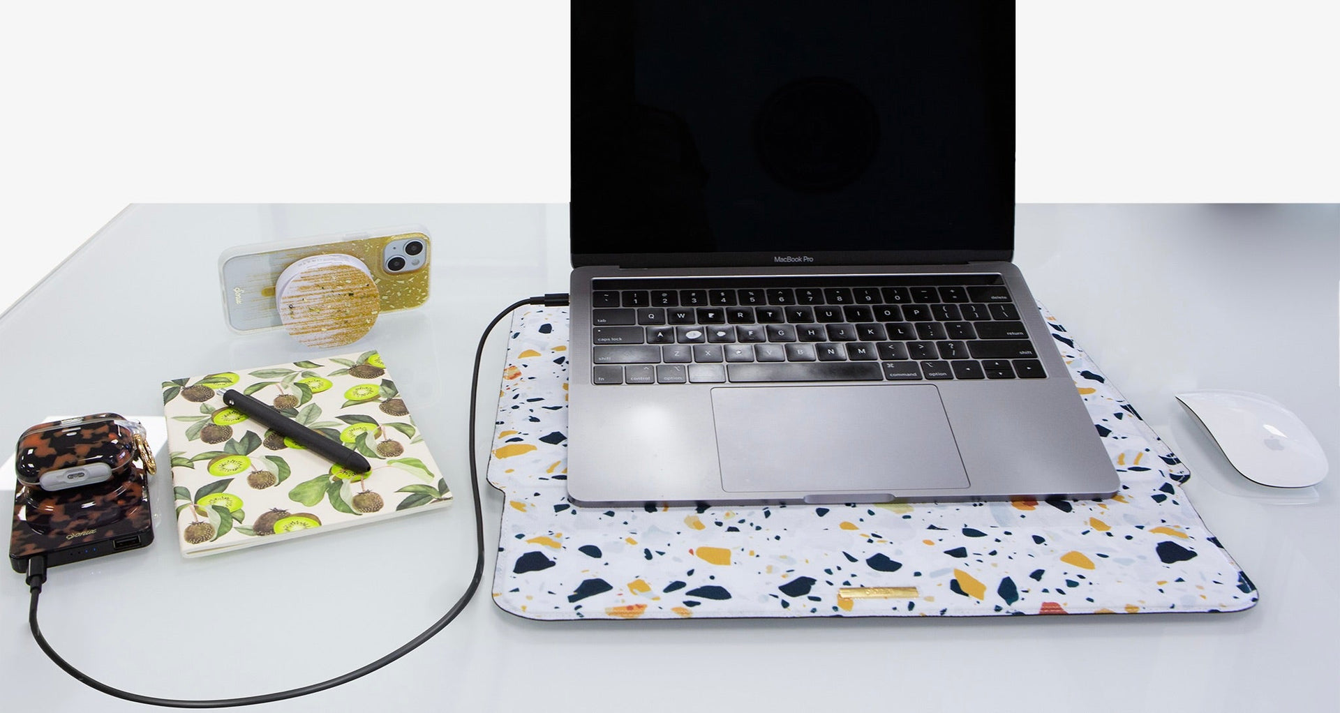 MacBook Pro on terrazzo laptop sleeve with wireless mouse, phone with gold PopSocket, and AirPods case