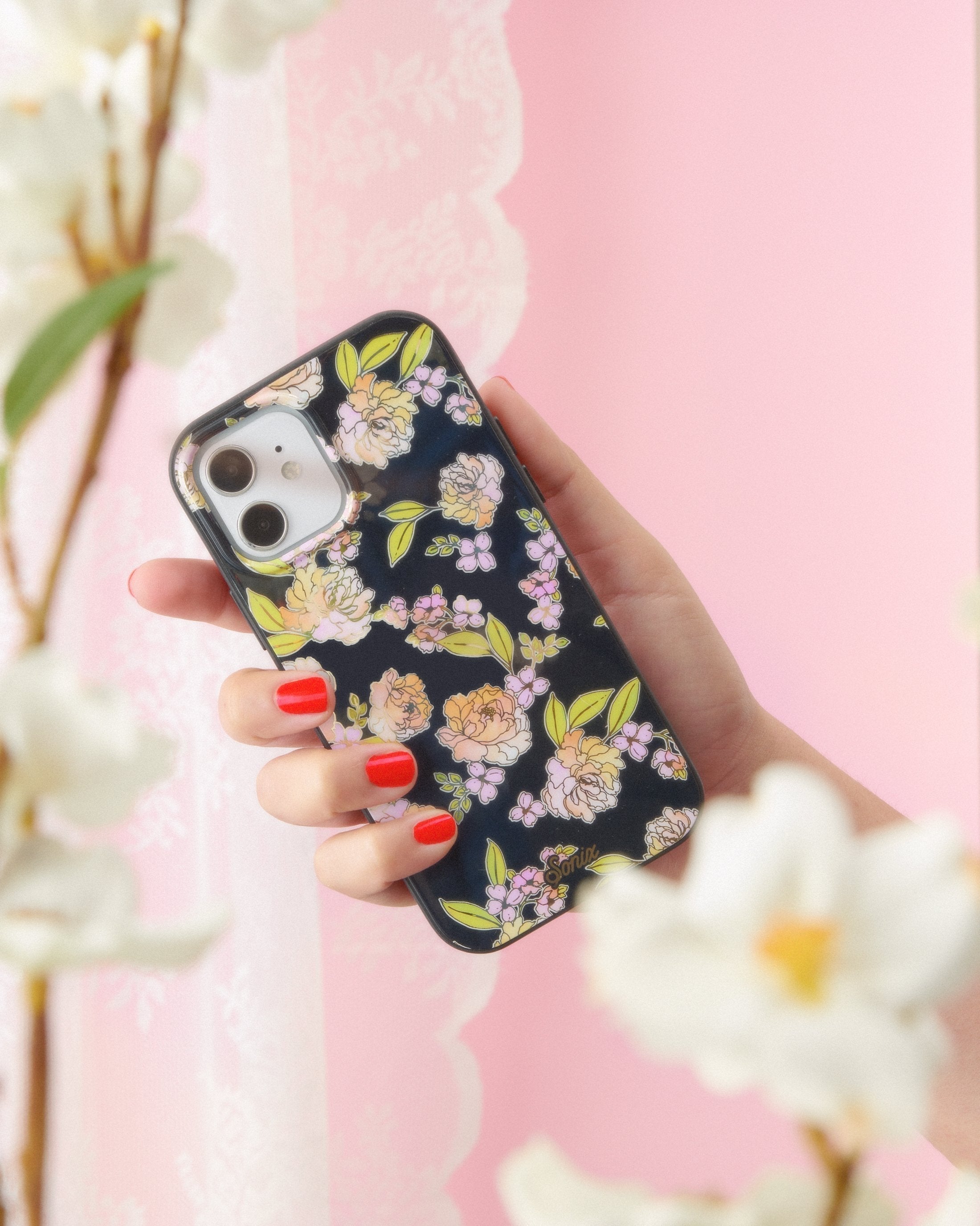 Black, iPhone case features a black base, gold foil details, and dainty flowers held by a hand with a pink background