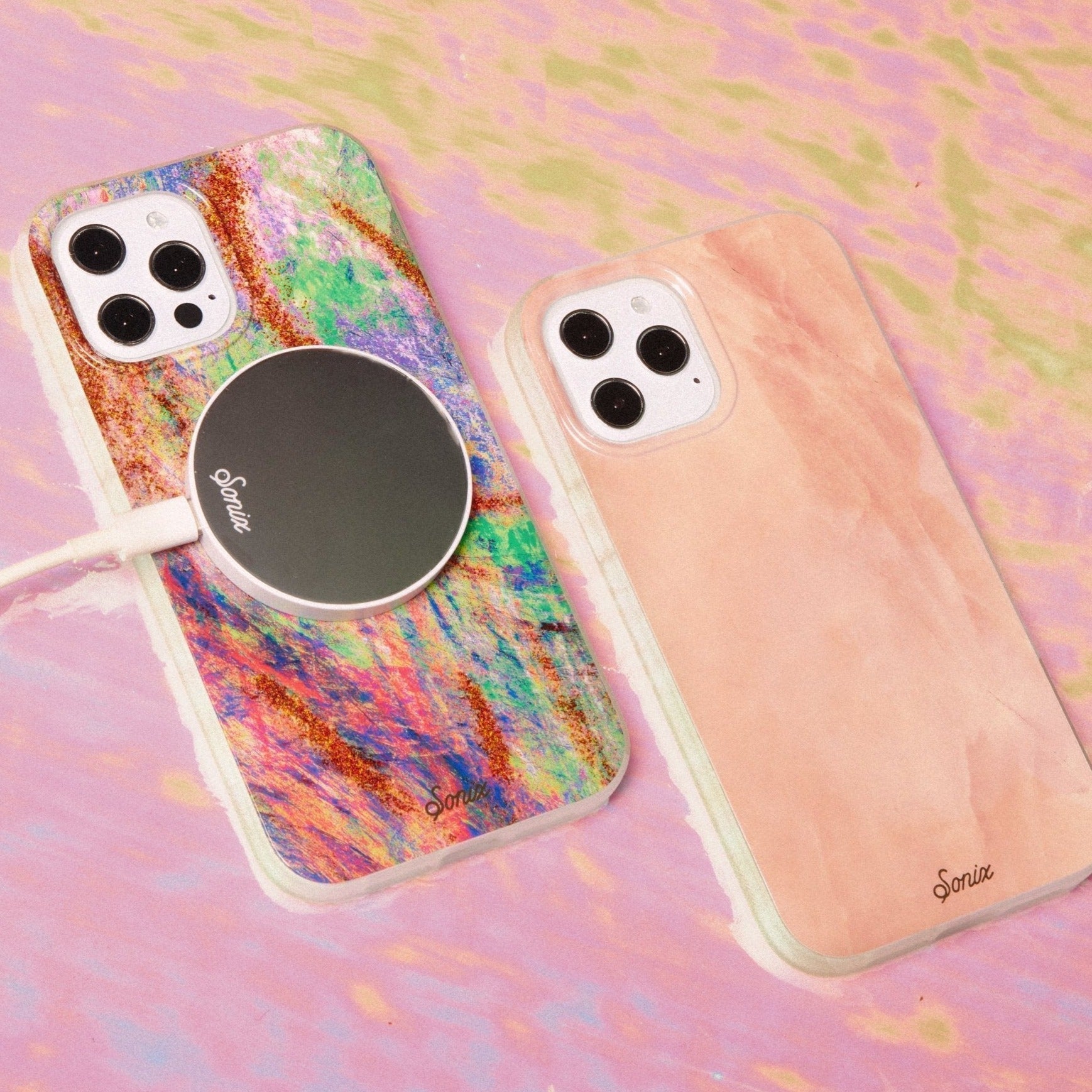  a paint splash of colors, resembling the Mardi Gras carnival, and finished with accents of gold glitter shown on an iphone with a magnetic link charger attached to the back and next to a pink mother of pearl phone case